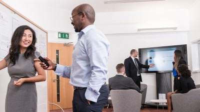 Male and female talking in front of a whiteboard with people looking at a tv screen in the background.