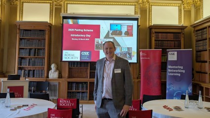 Prof Collomosse standing in a pannelled room, surrounded by baners, smiling.
