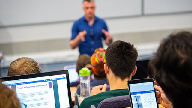 Biochemistry students are sitting a lecture, their laptops are open and a lecturer is presenting