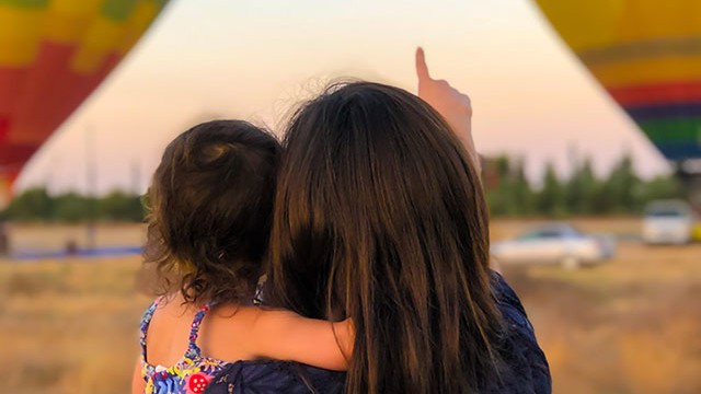 An adult is pointing at a hot air balloon while holding a child