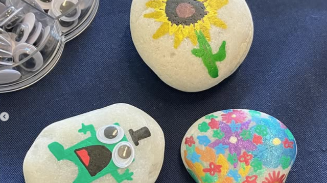 Three painted pebbles, one with a sunflower, one with multicoloured flowers, and one with a frog with googly eyes and a top hat