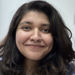An Indian woman with wavy black hair is wearing a nose ring and smiling softly at the camera.