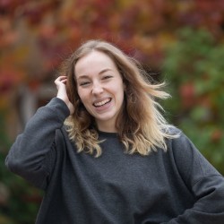 A young woman with light brown, wavy hair smiles brightly while standing outdoors. She is wearing a dark grey sweatshirt and holding one hand up to her hair. The background is softly blurred with autumn-coloured foliage in red, orange, and green hues.