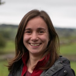 Picture of young woman with brown hair smiling against a rural background, wearing a red fleece and raincoat