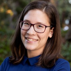 The image displays Zsofia Zavecz, who has long brown hair and wears glasses, smiling in a blue collared shirt.