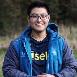 The researcher is pictured outdoors during fieldwork, wearing glasses, a blue jacket, and a dark sports jersey, smiling in a natural setting.