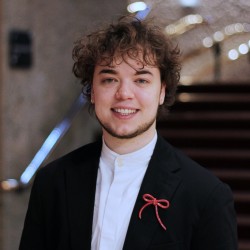 A photo of Kar Balan who is smiling and looking at the camera, in a formal setting, dressed in a black tuxedo and wearing a red bow broach.