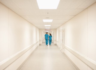 Two people wearing green scrubs can be seen a long way down a bright, white hospital corridor