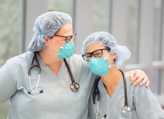 Two female, health care providers embrace while wearing light green medical scrubs, stethoscopes and N95 masks in an indoor hallway