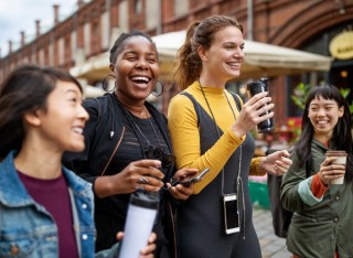 Four young women of different ethnic backgrounds enjoy a city break together. They are carrying phones and hot drinks and look as though they are having fun