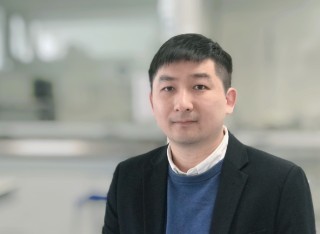 An Asian man wearing a dark jacket, blue jumper and white collared shirt stands in a light, airy building