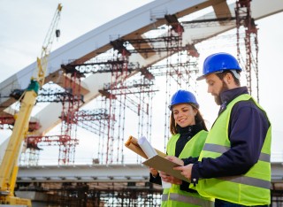 Two engineers, male and female, in protective helmets and reflective clothing, standing on construction site where large arch bridge is being built.
