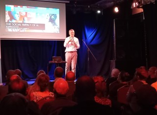 Andrew Rogoyski speaking at Pint of SDcience, screen behind him, audience's back of heads showing, watching him.