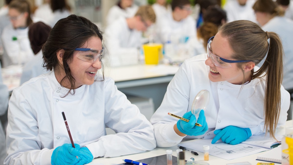 Two students in lab coats laughing, holding a petri dish